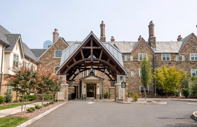 Front exterior view of a senior living facility with a large covered entrance supported by wooden beams, stone walls, multiple chimneys, and landscaped greenery including trees and shrubs.