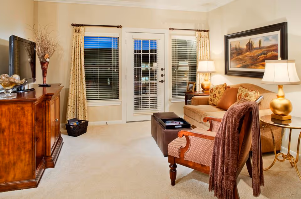 A cozy living room with beige walls and carpet. The room features a wooden TV stand with a flat-screen TV and decorative vase, two windows and a glass door with white blinds and patterned curtains. There is a beige sofa with patterned cushions, a wooden armchair draped with a brown throw, a brown ottoman, two side tables with golden lamps, and a framed landscape painting on the wall.