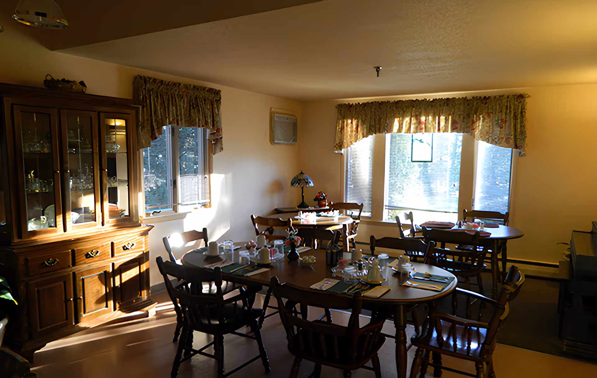 Sunlit dining room with round wooden tables and chairs set for a meal and a china cabinet by the window.