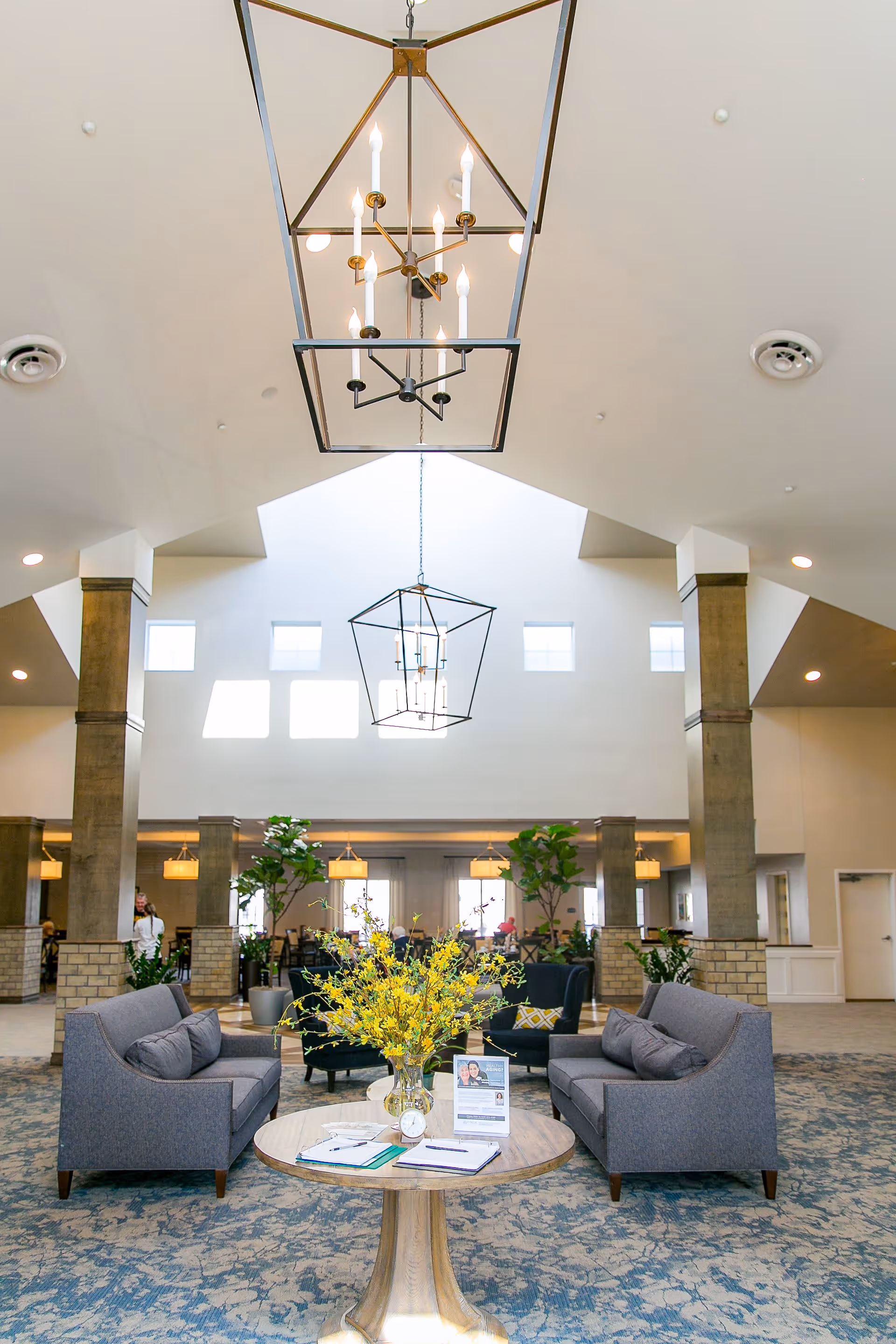 A spacious and well-lit senior living facility common area with high ceilings and large windows. The room features two gray sofas facing each other with a round wooden table in between, decorated with a vase of yellow flowers and informational brochures. There are large hanging light fixtures and several potted plants around the seating area, creating a welcoming atmosphere.