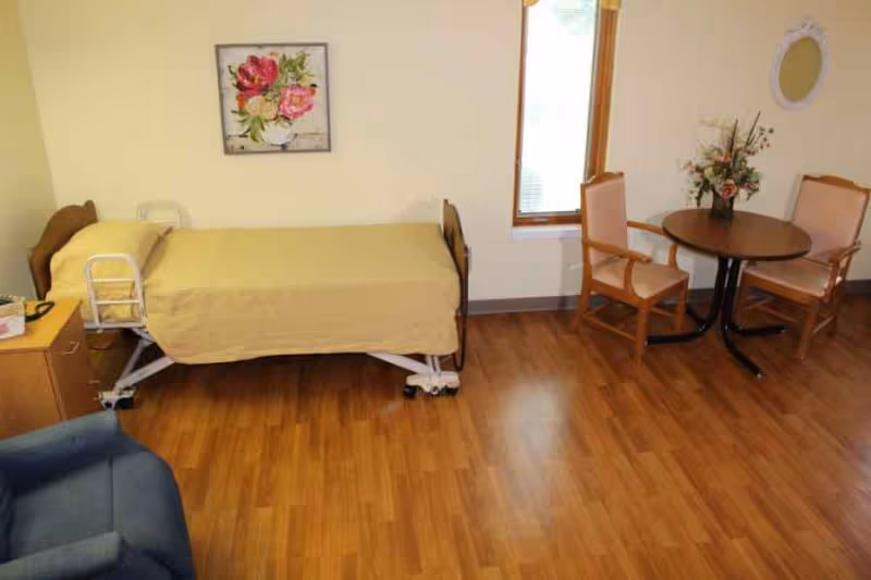 Nursing-home bedroom with a single hospital bed, small round table with two chairs, and hardwood flooring.
