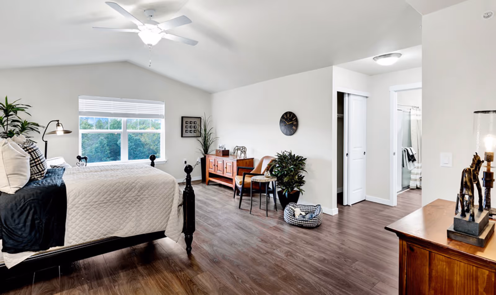 A spacious, well-lit bedroom with a large window showing greenery outside. The room features a bed with white and black bedding, a ceiling fan with light, wooden flooring, a wooden dresser with decorative items including a lamp and giraffe figurines, a small wooden cabinet, a leather chair, a side table, a potted plant, a wall clock, and an open doorway leading to a bathroom and closet.