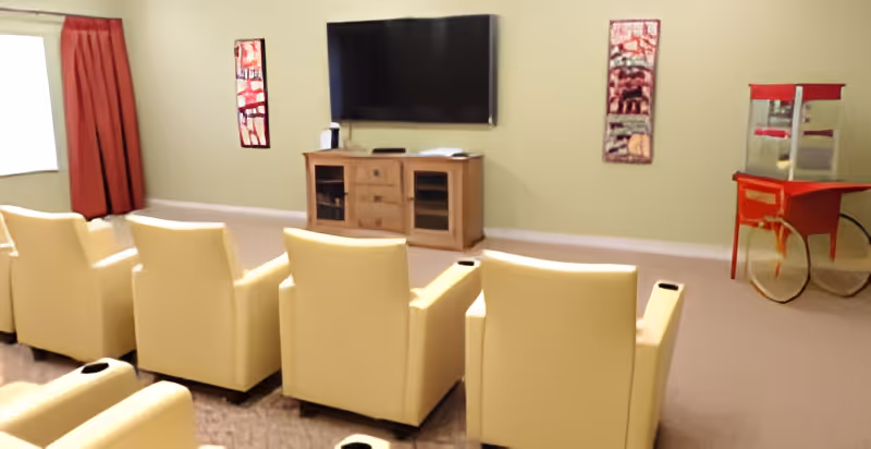 Row of yellow recliner chairs facing a wall-mounted TV and media console in a common lounge with a popcorn machine.