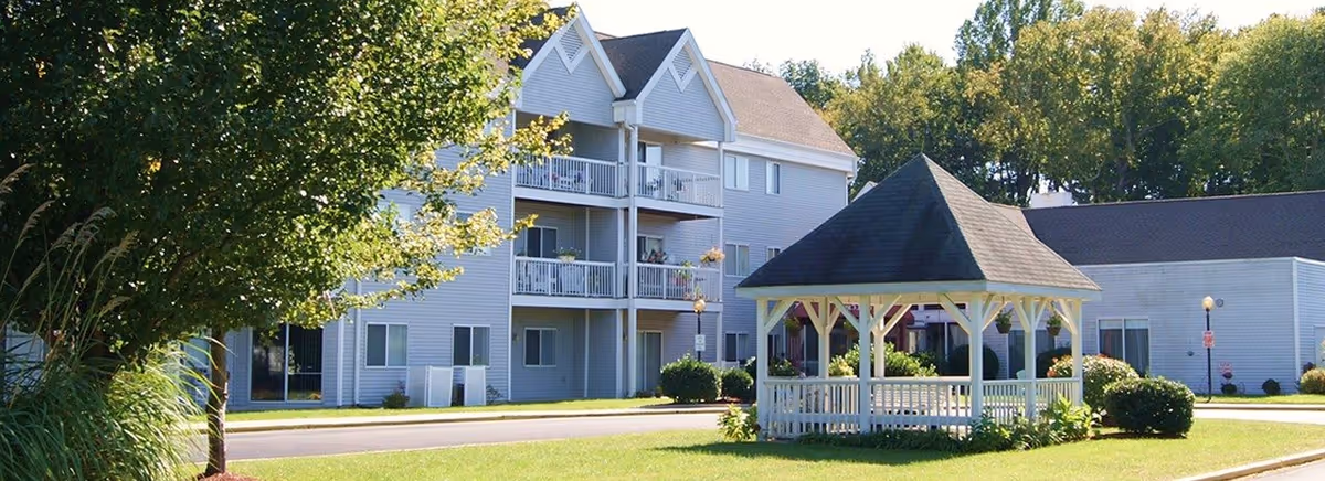 Outdoor view of a senior living facility with a white gazebo on a grassy area, surrounded by bushes and trees. In the background, there is a multi-story building with balconies and windows.