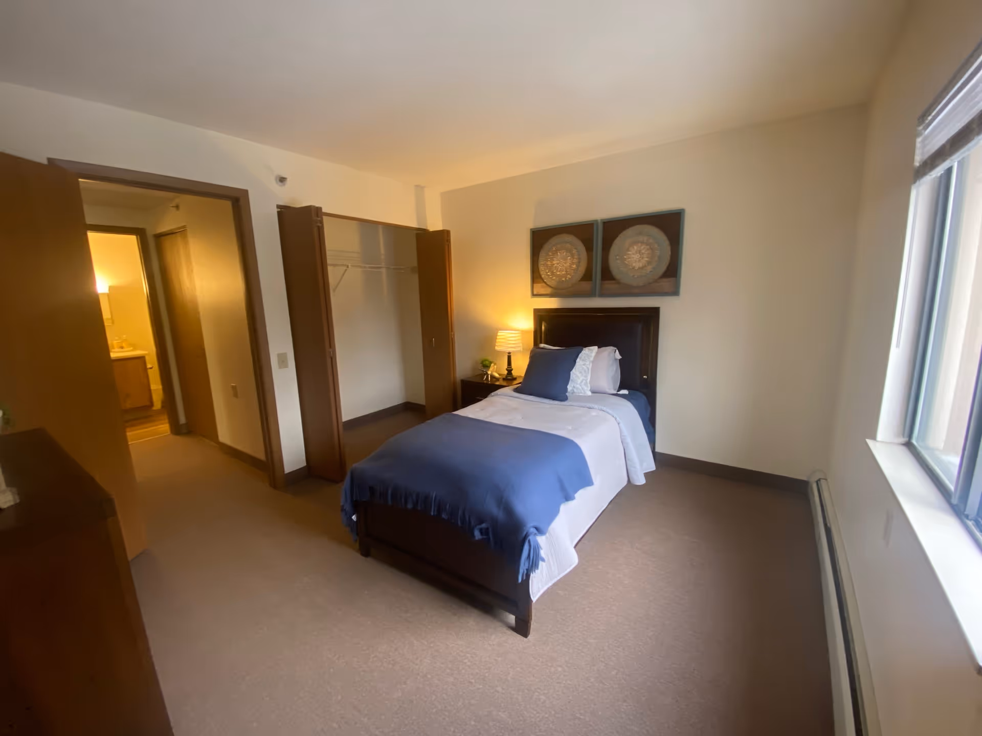 A furnished bedroom with a single bed dressed in blue bedding, a nightstand with a lamp, an open closet, and a window.