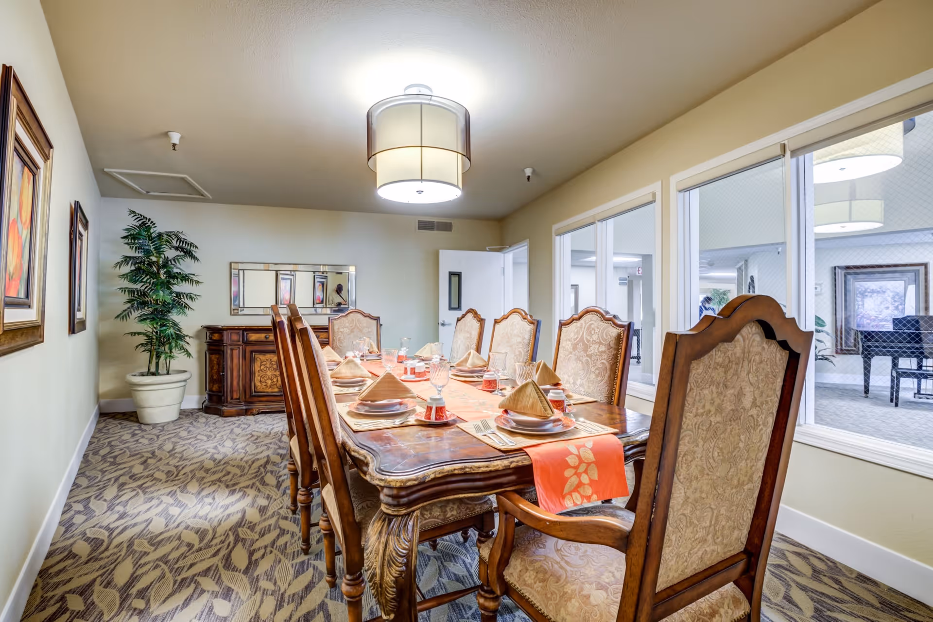 A formal dining room with a long wooden table set for eight people. The table is adorned with folded napkins, plates, glasses, and small red cups. The chairs have ornate wooden frames and upholstered backs and seats. The room has patterned carpet, light-colored walls, a large ceiling light fixture, a potted plant in the corner, framed artwork on the wall, and a sideboard with a mirror above it. Large windows on one side look into an adjacent room with a piano and additional seating.