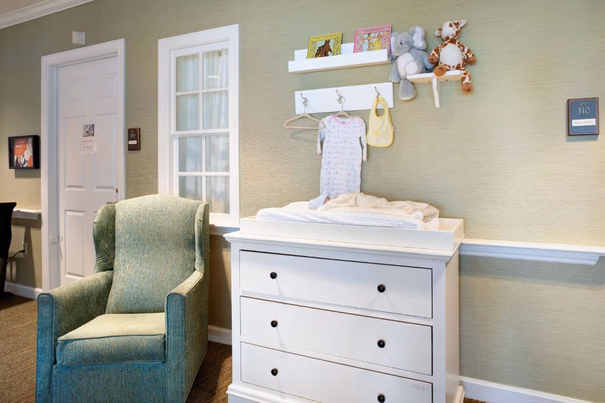 A cozy corner of a room featuring a green upholstered armchair next to a white dresser with three drawers. On top of the dresser is a changing pad with folded blankets. Above the dresser, a wall-mounted shelf holds children's books and stuffed animals, with a baby outfit and bib hanging on hooks beneath the shelf. The walls are light green with white trim, and there is a white door and window in the background.