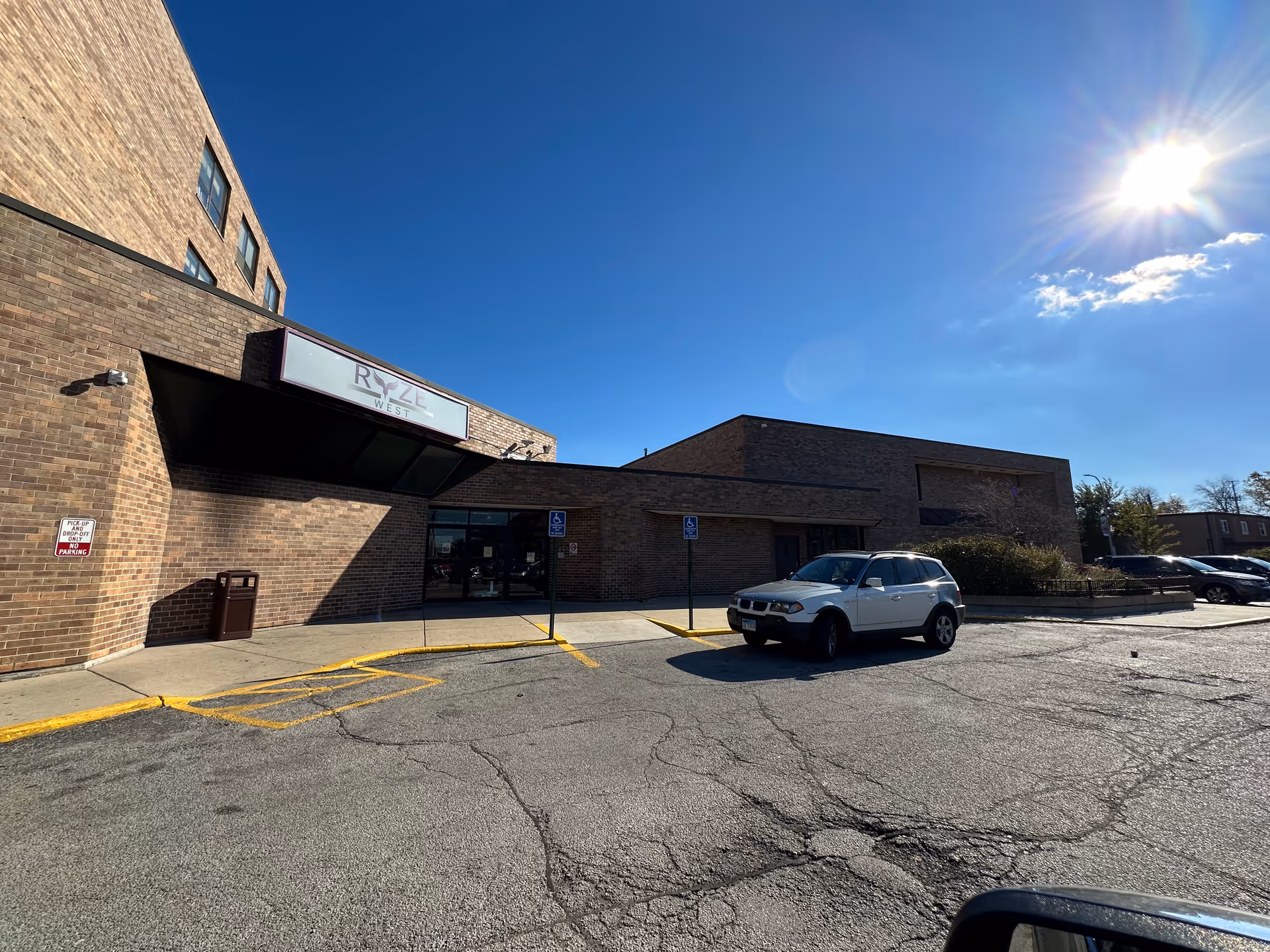 Exterior view of a brick building under a clear blue sky with the sun shining brightly. The building has a sign that reads 'RYZE WEST' above the entrance. There is a parking lot in front with a white SUV parked near the entrance and two handicap parking signs visible.