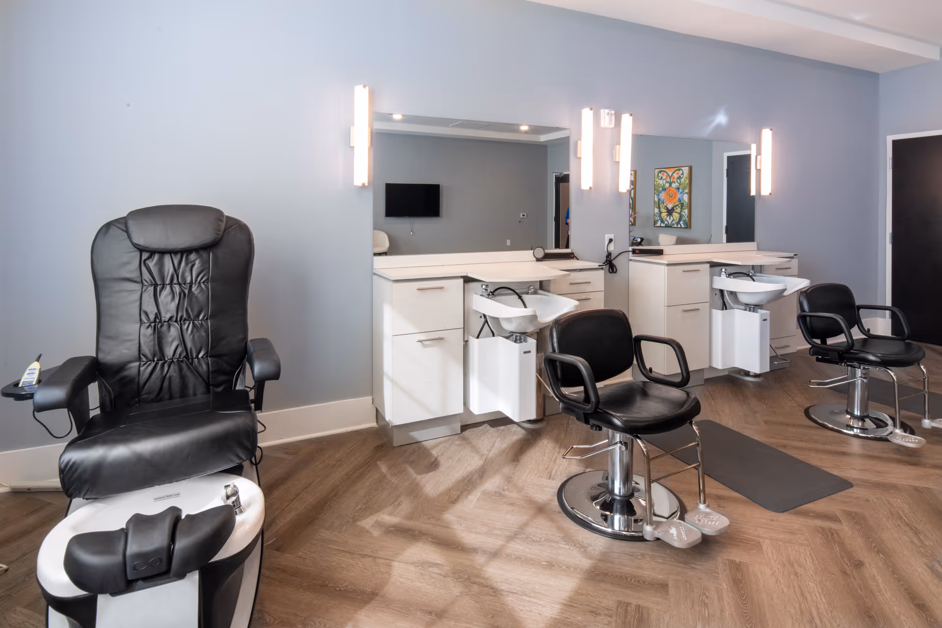 Interior of a salon area in a senior living facility featuring a black leather pedicure chair with foot bath on the left, two black salon chairs in front of white cabinetry with sinks and large mirrors, soft wall lighting, and a wooden floor.