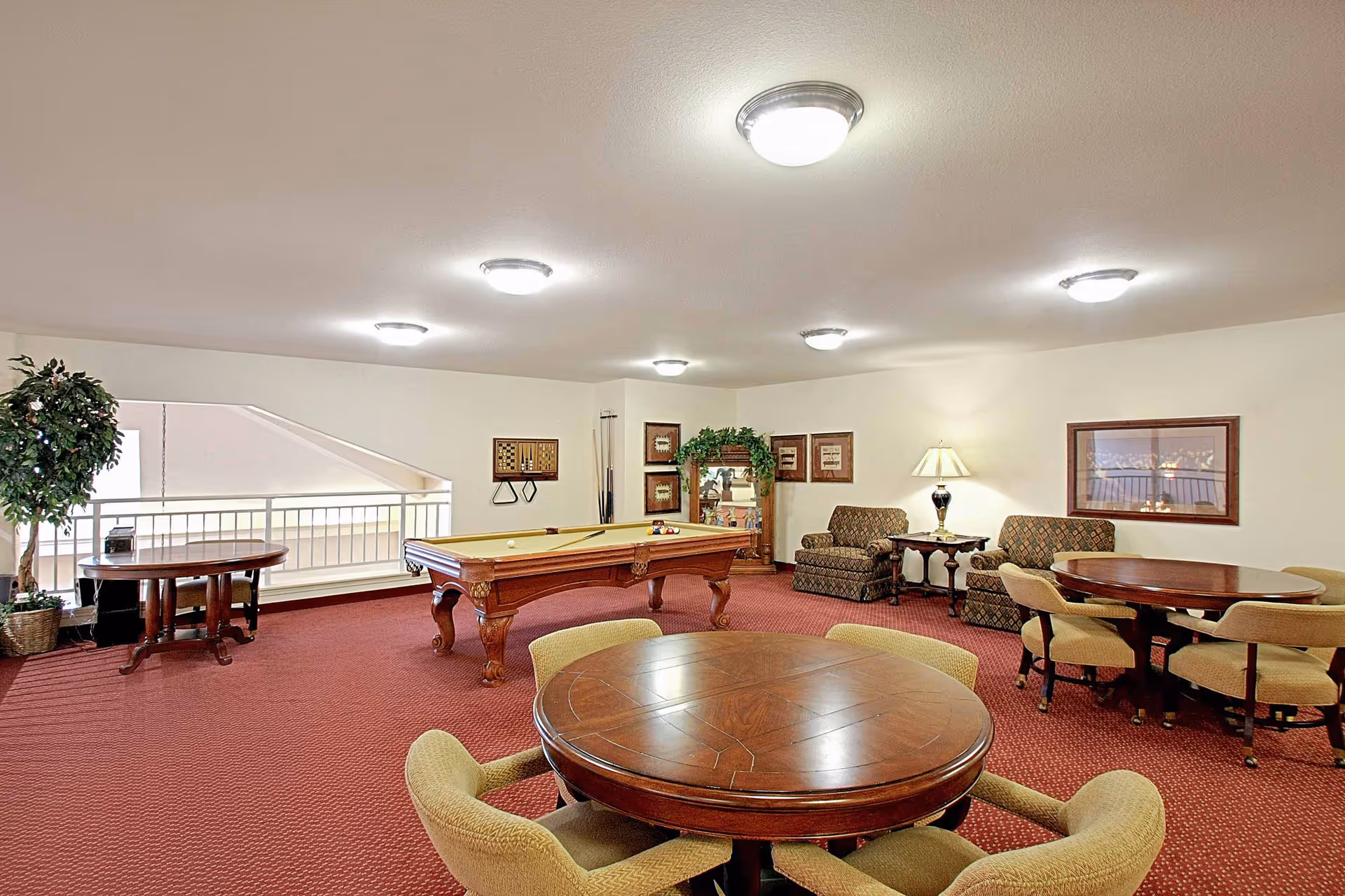 A senior living facility game room with a pool table in the center, surrounded by round wooden tables with cushioned chairs. There are two upholstered armchairs with a side table and lamp against the wall. The room has red carpet flooring, white walls, framed artwork, and ceiling lights. A potted plant is near a railing overlooking a lower level.