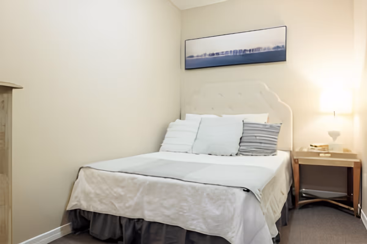 A small bedroom with a neatly made bed featuring a tufted beige headboard, several pillows in white and gray tones, a light-colored blanket, a wooden nightstand with a lamp, and a framed landscape artwork hanging above the bed.