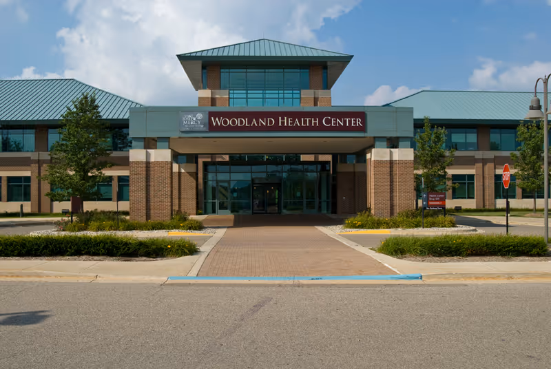 Front entrance of a brick health center with a covered drive-up, teal metal roof, and a sign reading "Woodland Health Center" under a partly cloudy sky.