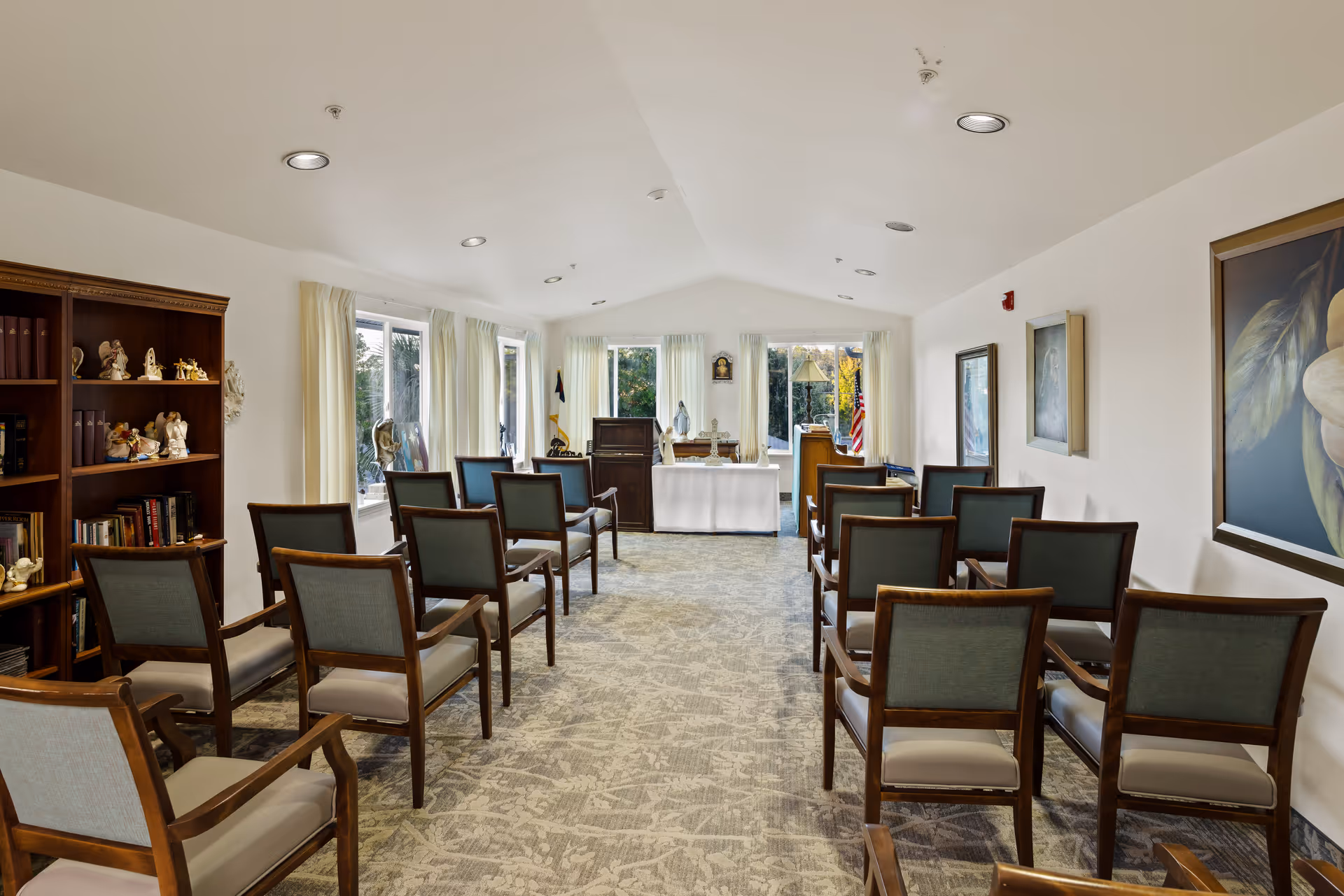 A small chapel or meditation room with rows of wooden chairs facing an altar at the front. The room has large windows with white curtains letting in natural light, a bookshelf with decorative items on the left, and framed artwork on the right wall.