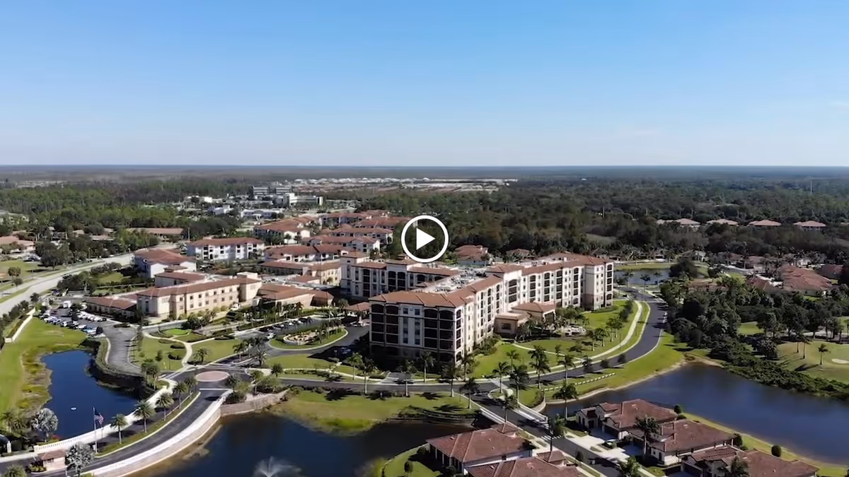 Aerial view of The Arlington of Naples senior living facility showing multiple connected buildings with red-tiled roofs surrounded by landscaped greenery, water features, and roads under a clear blue sky.