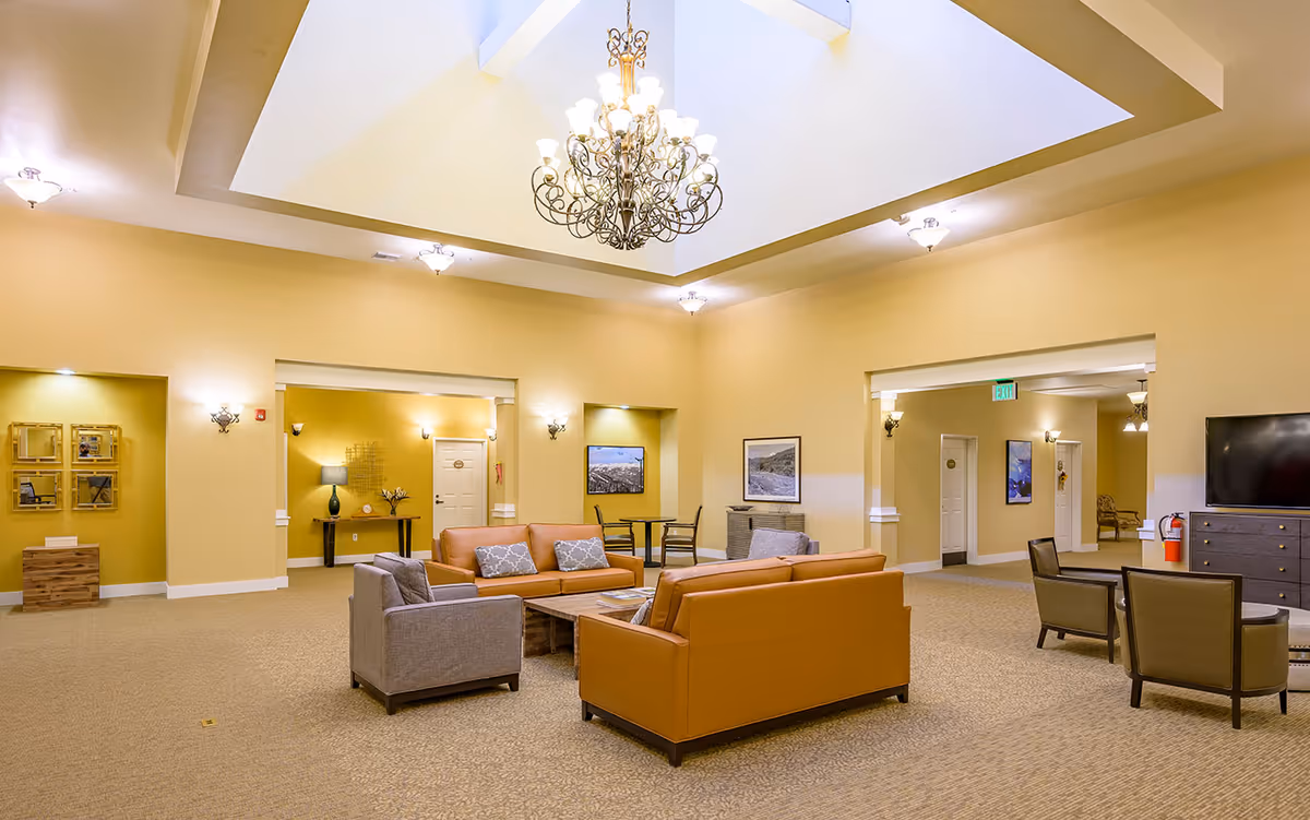 A spacious and well-lit common area in an assisted living facility featuring beige walls and carpeted floors. The room has a large decorative chandelier hanging from a recessed ceiling. There are two brown leather sofas and two gray armchairs arranged around a wooden coffee table in the center. Additional seating includes two chairs near a wall-mounted TV on a dark wooden dresser. The room has multiple doorways leading to other areas, with framed pictures on the walls and soft lighting fixtures.