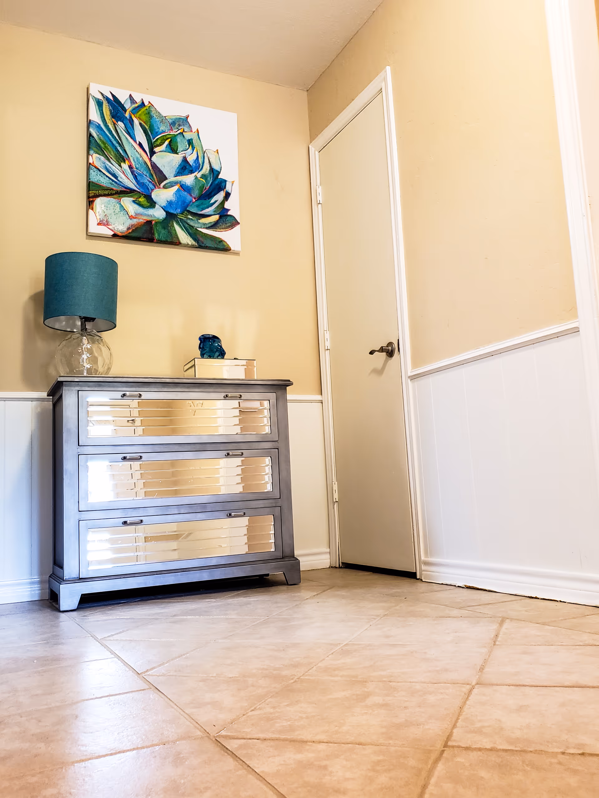 A corner of a room with beige walls and tiled floor, featuring a mirrored three-drawer chest with a glass lamp with a teal lampshade and a small decorative item on top. Above the chest hangs a colorful painting of a succulent plant. A closed white door with a black handle is visible to the right.