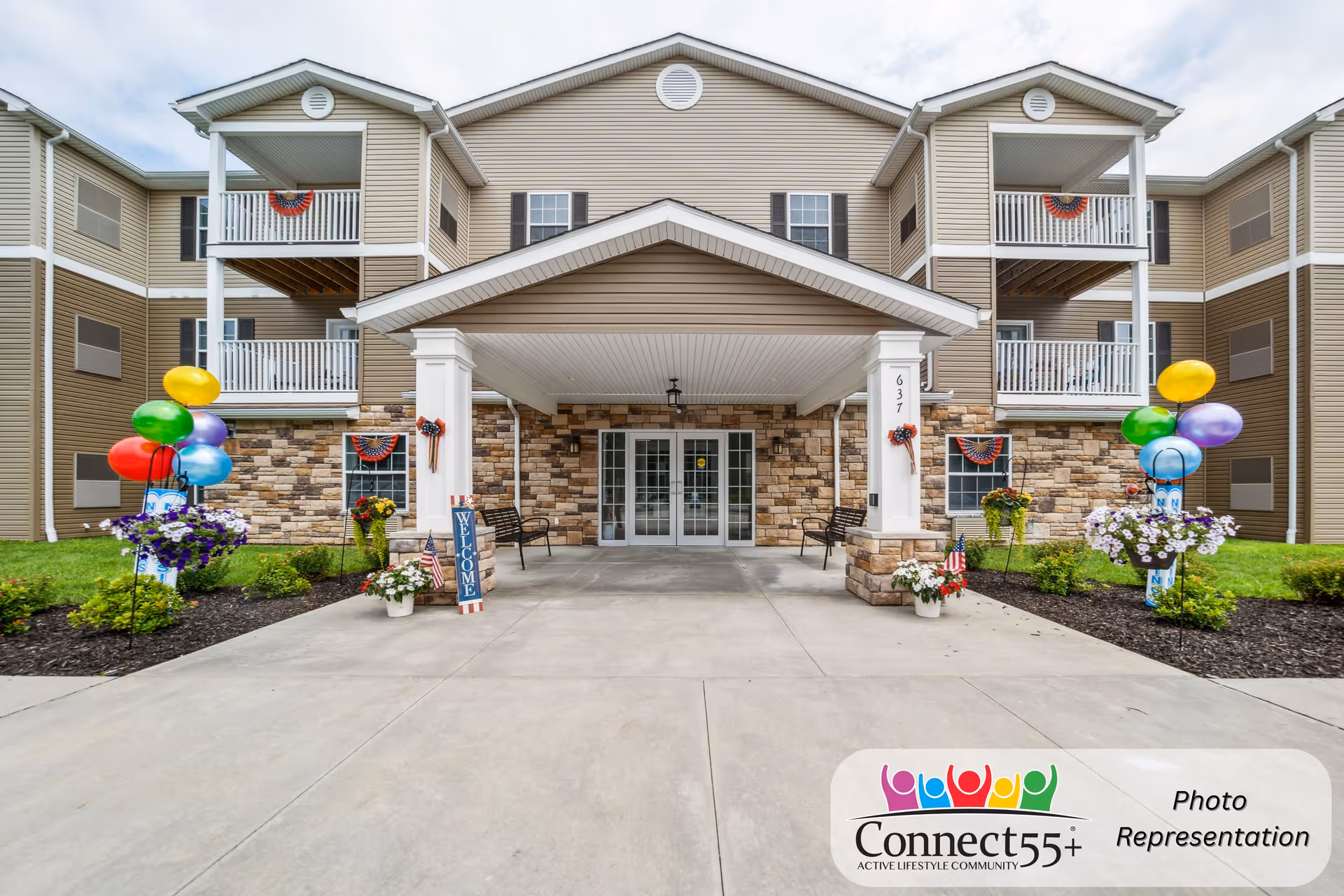 Front exterior view of a senior living community building with a covered entrance, stone and beige siding facade, balconies, colorful balloons, flower pots, and patriotic decorations.