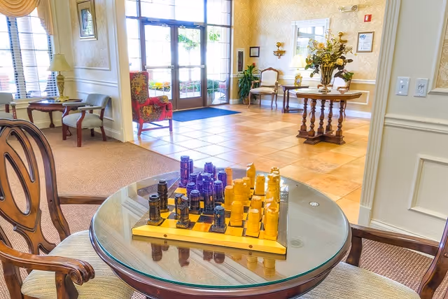 A bright and welcoming senior living facility common area with a glass-top round table featuring a chessboard and chess pieces. The room has comfortable wooden chairs, a tiled floor, large windows, and a glass door leading outside. There are decorative plants, a round wooden table with a floral arrangement, and framed artwork on the walls.