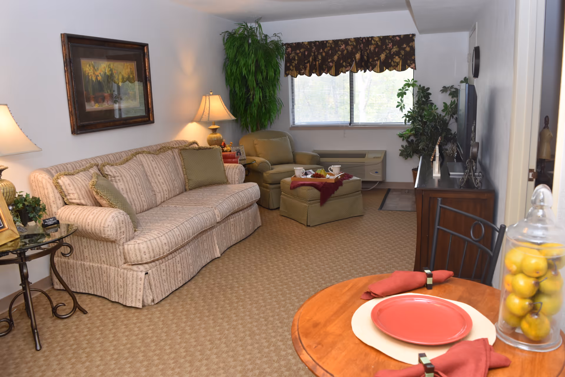 Well-lit living room with a striped sofa, armchair and ottoman, TV cabinet, and a small dining table set with plates and napkins.