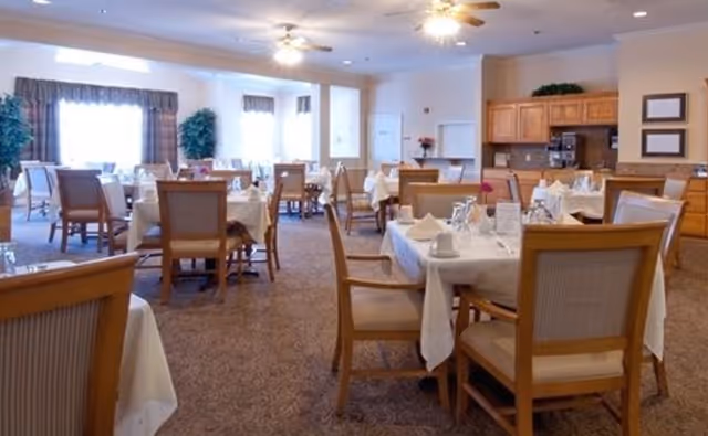 Bright dining room with multiple set tables and wooden chairs in a senior living facility.