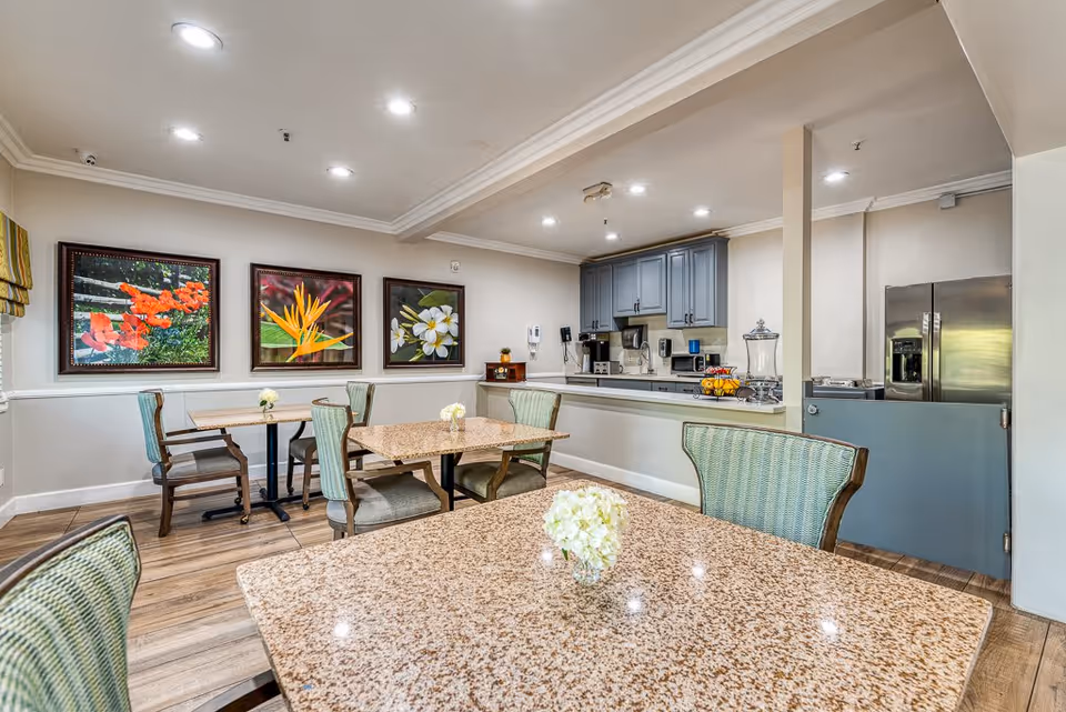 A bright and clean dining area in a senior living facility with several tables and chairs. The tables have a speckled granite surface and small floral centerpieces. The room features a kitchenette with gray cabinets, a stainless steel refrigerator, and various appliances. Three framed floral paintings hang on the wall, and the floor is wood-patterned.