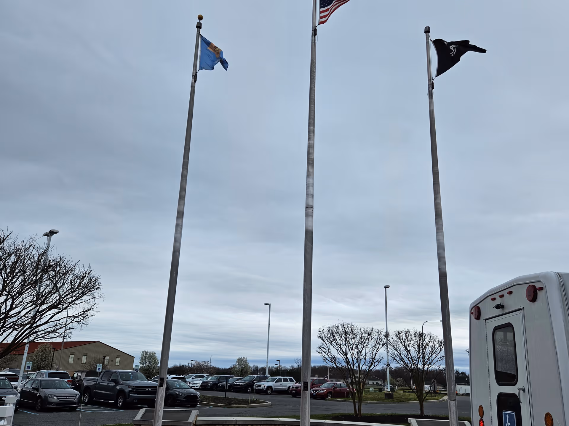 Three flagpoles with flags flying against a cloudy sky in a parking lot area with several parked cars and leafless trees. A white vehicle is partially visible on the right side.