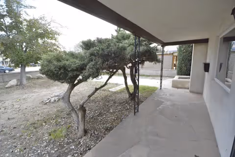 Covered front porch and walkway of a single-story building overlooking a sparse yard with low, wind-sculpted trees.
