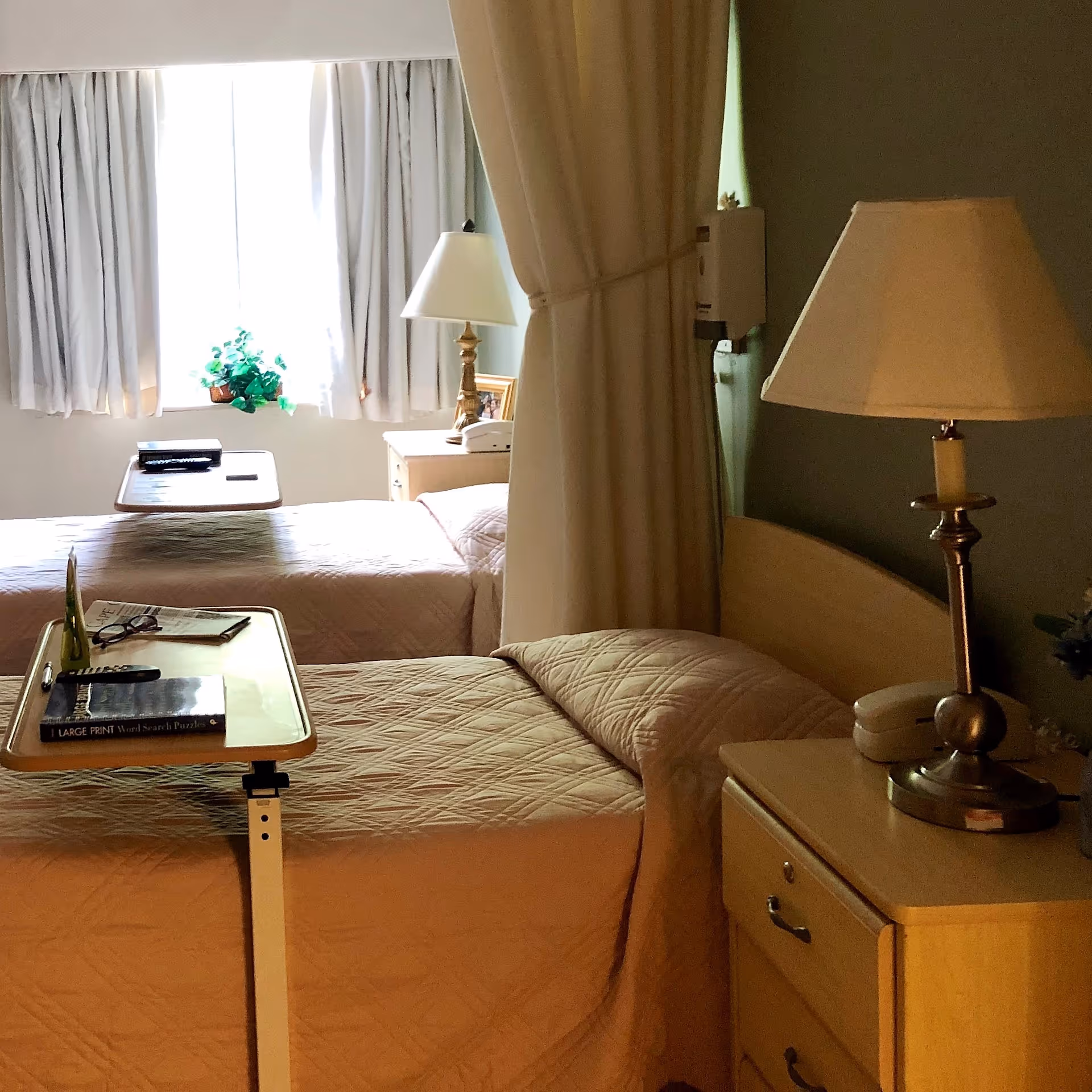 A shared bedroom in a senior living facility with two beds separated by a curtain. Each bed has a beige quilted bedspread, a bedside table with a lamp, and a small overbed table with items such as books, glasses, and a remote control. A window with white curtains allows natural light into the room.