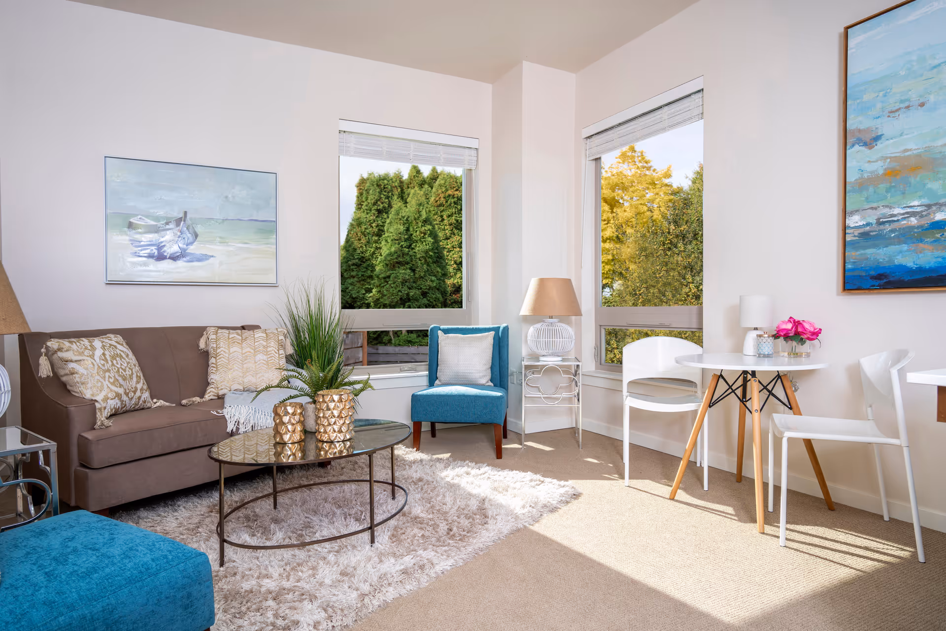 A bright and cozy living room with a brown sofa adorned with patterned pillows, a blue armchair, a glass coffee table with decorative plants, a white round dining table with two white chairs, and large windows showing green trees outside. The room features light-colored walls, a fluffy beige rug, and colorful artwork on the walls.