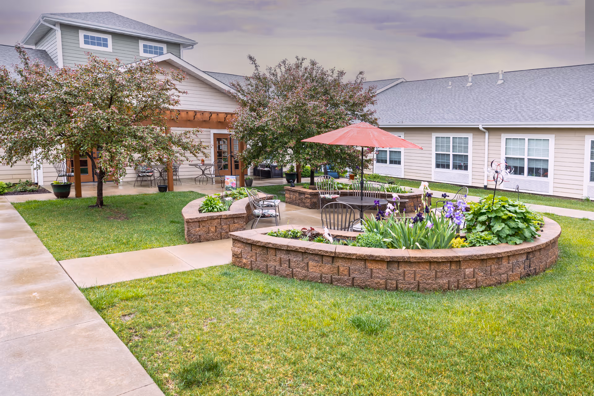 Outdoor courtyard area at Oak Pointe of Kearney featuring a circular raised garden bed with flowers and plants, metal chairs, a table with a red umbrella, and trees with pink blossoms. The courtyard is surrounded by beige buildings with multiple windows and a covered patio area with additional seating.