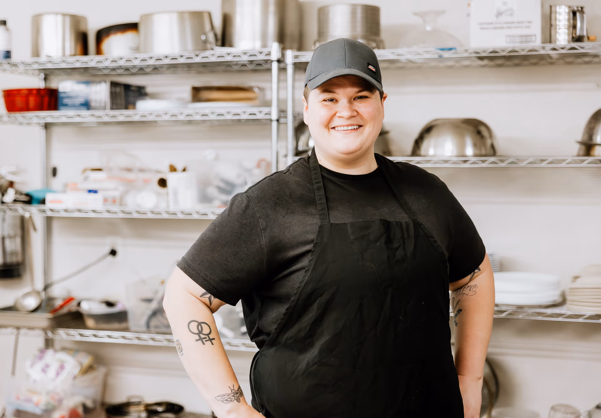 A smiling person wearing a black apron and a gray cap stands in a kitchen with metal shelves filled with kitchenware and supplies in the background.