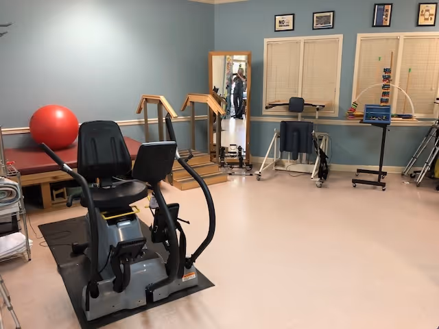 A physical therapy or rehabilitation room with exercise equipment including a recumbent exercise bike, parallel bars for walking practice, a large red exercise ball, a padded treatment table, a mirror, and various therapy tools and equipment against blue walls with framed pictures and windows with blinds.