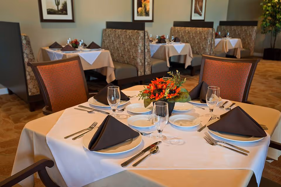 A dining table set for four with white and beige tablecloths, black folded napkins, plates, cutlery, and glasses. A small floral centerpiece with orange flowers is in the middle. In the background, there are booth seating areas with tables similarly set, framed pictures on the wall, and a potted plant in the corner.