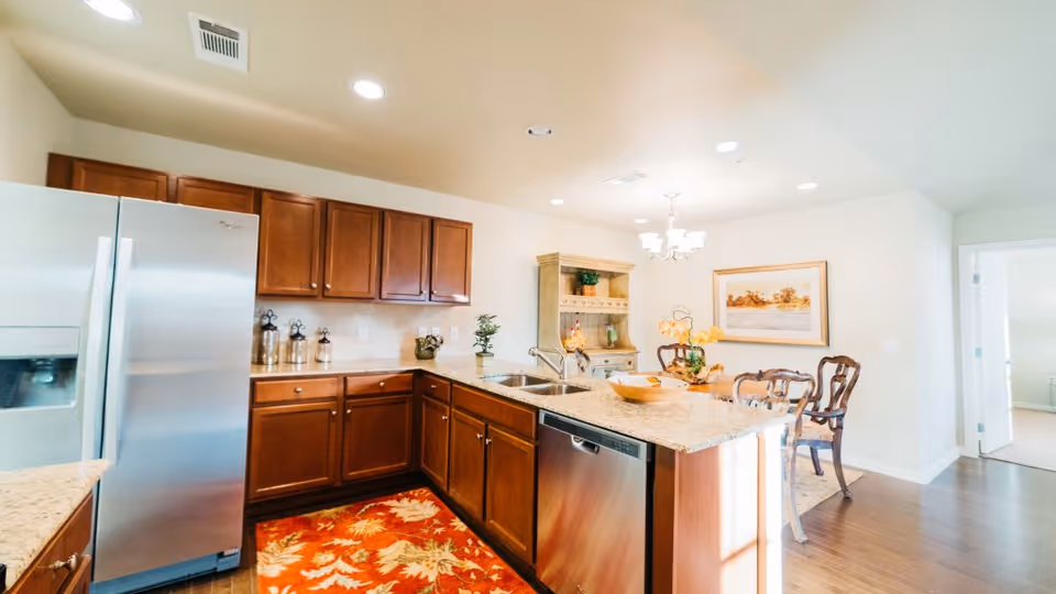 Bright kitchen area with wooden cabinets, stainless steel refrigerator and dishwasher, granite countertops, and a colorful rug on the floor. In the background, there is a dining area with a wooden table, chairs, a chandelier, and a framed picture on the wall.