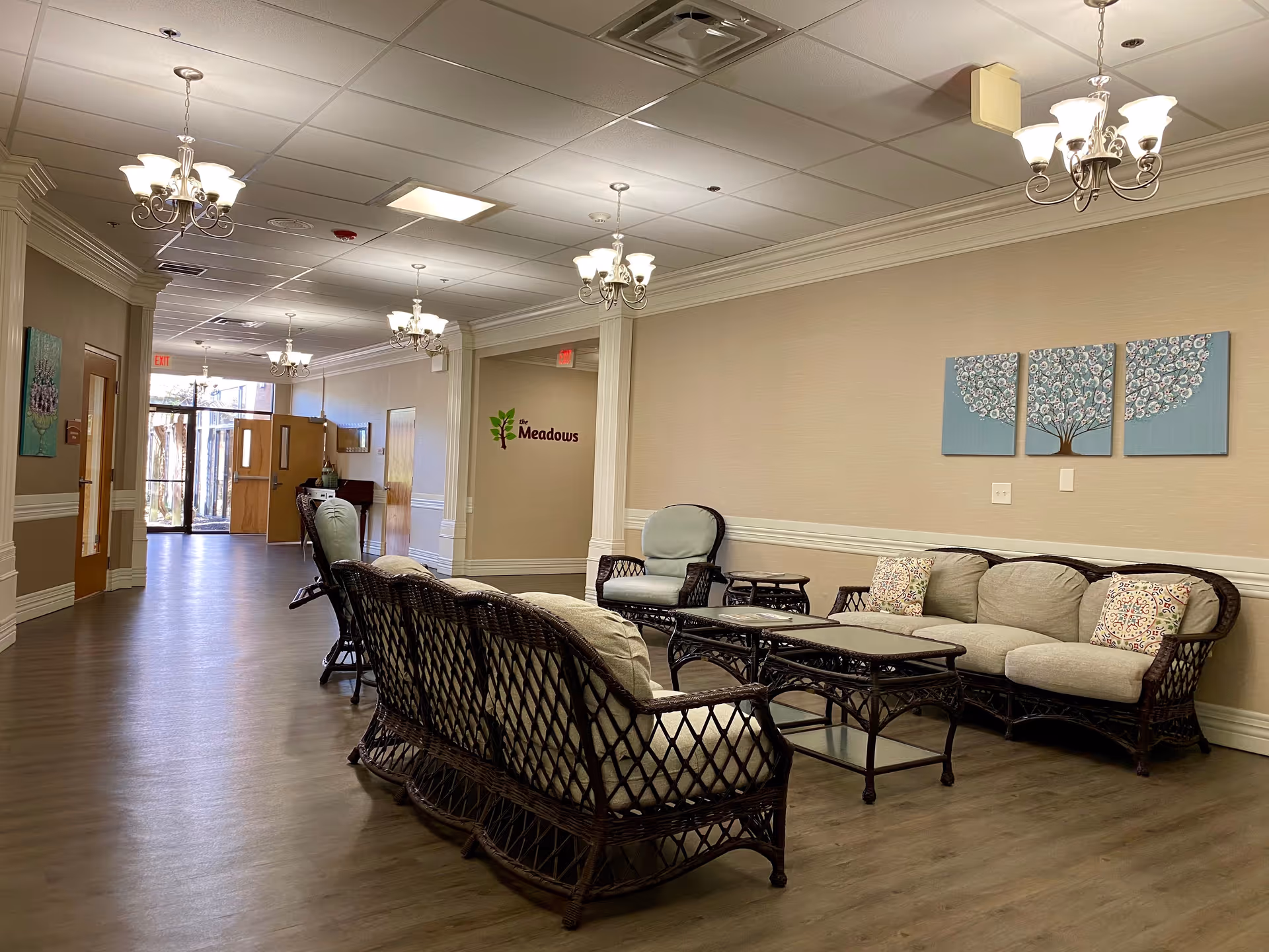 A well-lit hallway in a senior living facility with wicker seating furniture including sofas and chairs arranged along the right wall. The walls are beige with white trim, and there are multiple chandeliers hanging from the ceiling. A three-panel artwork of a tree is mounted on the wall above the sofa. The hallway extends to a glass door at the far end, and a sign on the wall reads 'The Meadows'.