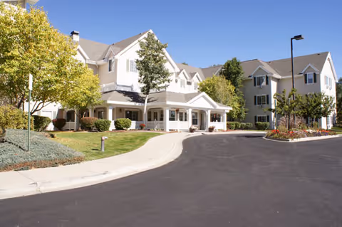 Front exterior of a multi-story senior living building with a covered entrance, curved driveway, and landscaped grounds under a clear blue sky.