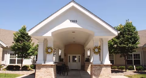 Front exterior view of a single-story building with a covered entrance supported by four white columns. The building has beige siding, a peaked roof, and the number 1365 displayed above the entrance. There are two trees and some landscaping on either side of the entrance.