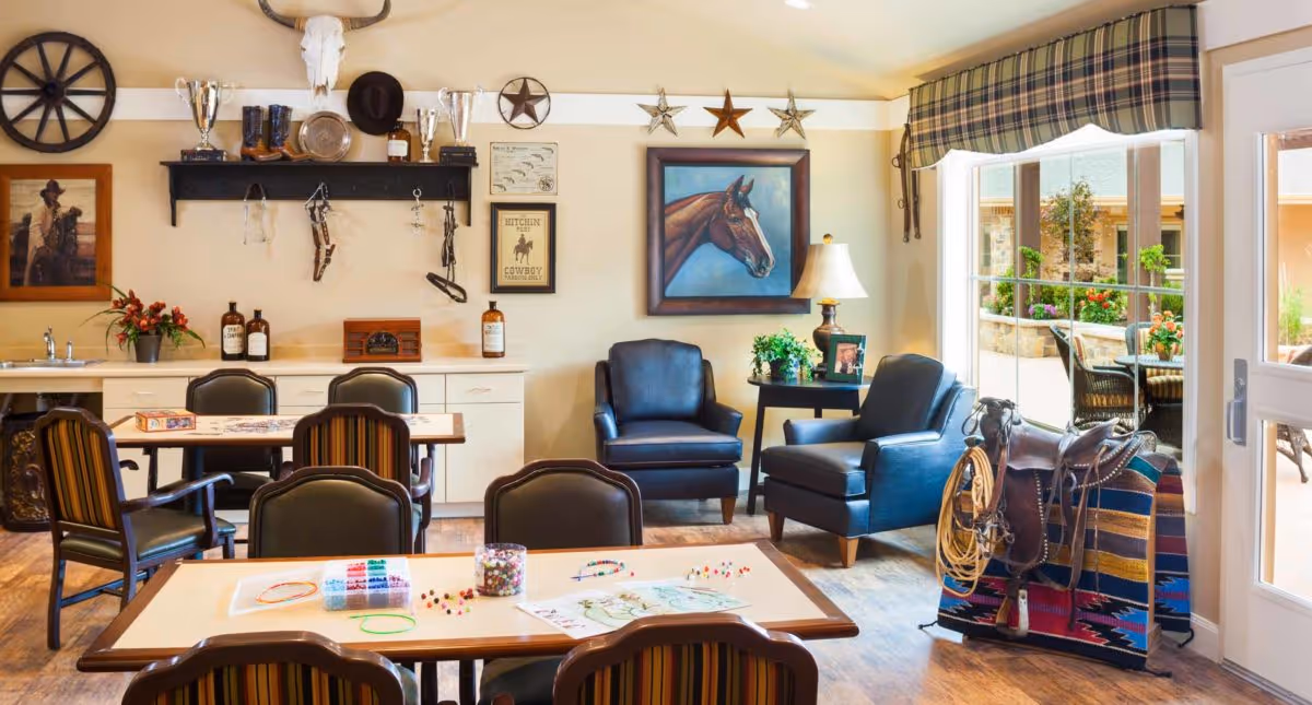 A cozy common area in Meadowbrook Memory Care Community featuring tables and chairs arranged for activities, two blue armchairs near a side table with a lamp and plant, western-themed decor including a horse painting, cowboy hats, boots, and a saddle, and large windows opening to an outdoor patio with seating and plants.