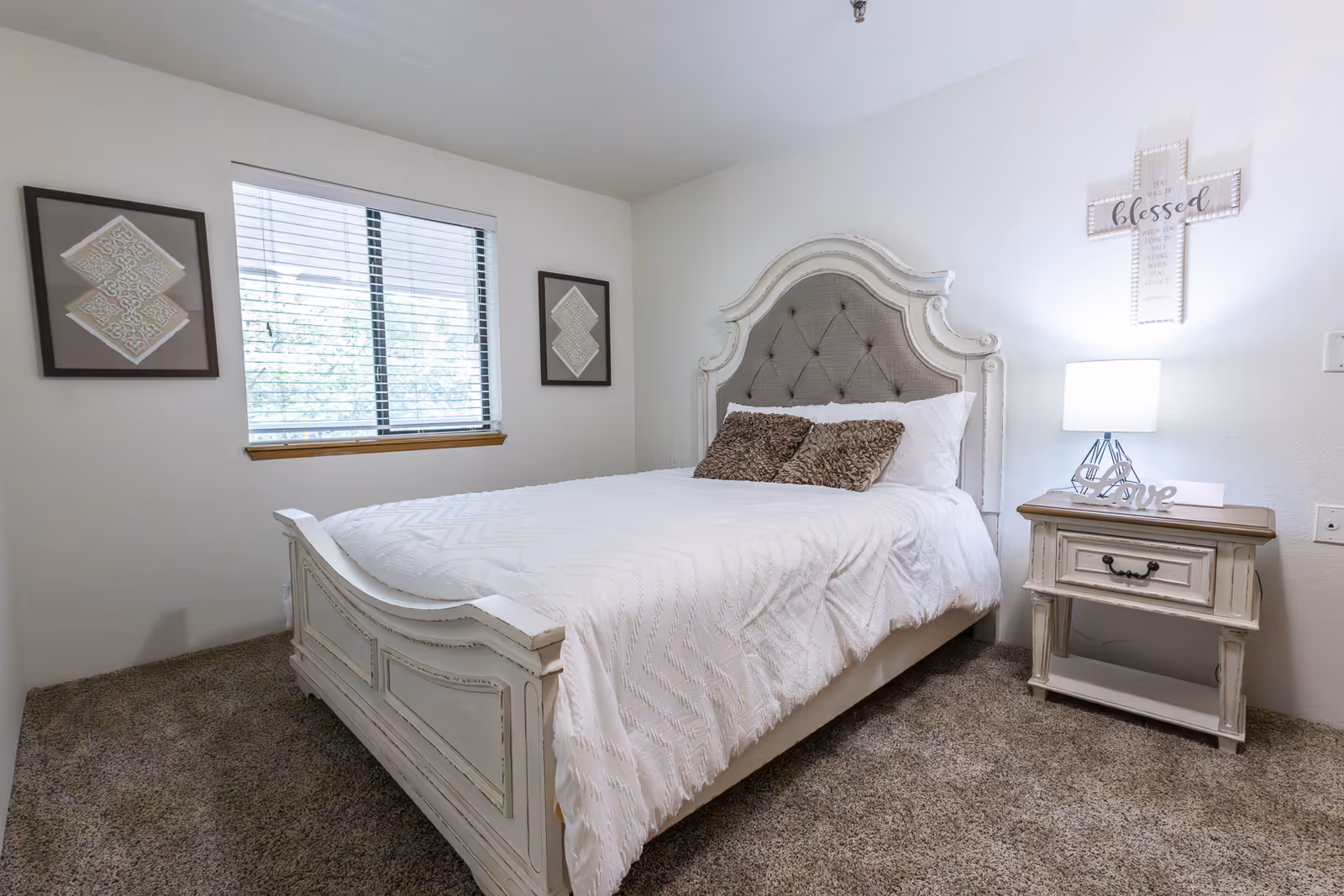 A cozy bedroom with a white ornate bed frame and a tufted headboard. The bed is made with white bedding and two brown textured pillows. There is a nightstand with a lamp and decorative items including a 'love' sign. On the wall above the nightstand is a cross-shaped decoration with the word 'blessed'. Two framed artworks hang on either side of a window with blinds. The room has beige carpet and white walls.