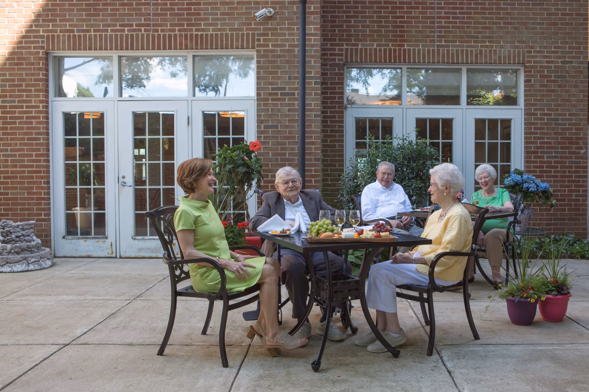 Four elderly people and one middle-aged woman sitting around two outdoor tables with food and drinks, enjoying a social gathering on a patio outside a brick building with large glass doors and windows.