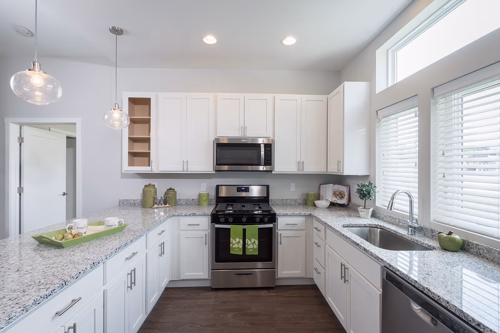 A modern kitchen with white cabinets, granite countertops, and stainless steel appliances including a stove and microwave. There are two pendant lights hanging from the ceiling, a sink under two large windows with blinds, and various decorative items on the counters including green towels hanging on the oven door.