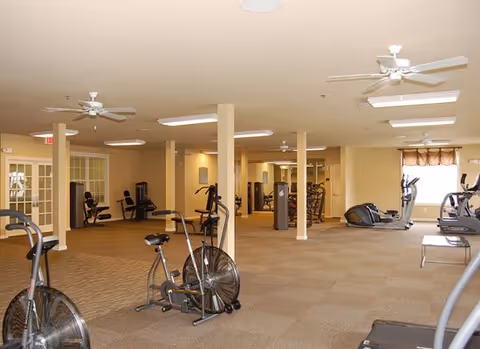 Spacious carpeted fitness room with exercise bikes, treadmills, and strength equipment under ceiling fans.