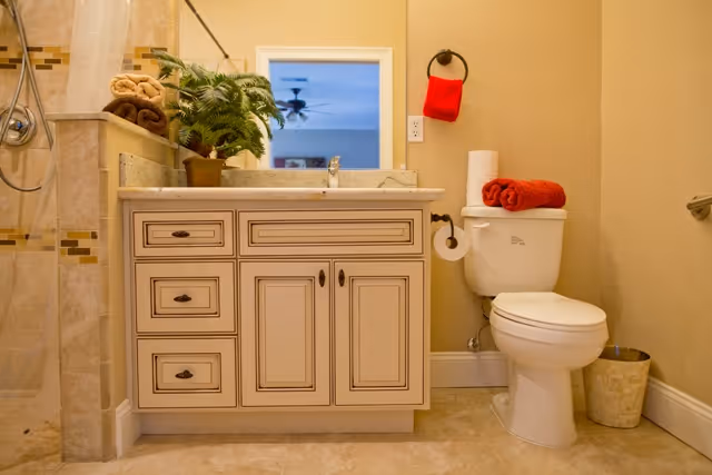 A well-lit bathroom featuring a vanity with sink and mirror, a toilet with folded towels on top, and a tiled shower to the left.