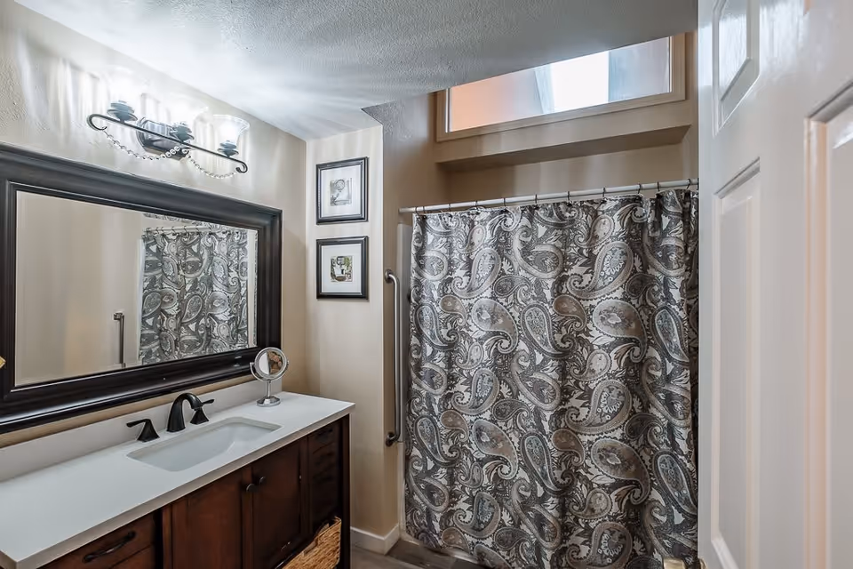 A bathroom with a dark wood vanity featuring a white countertop and an undermount sink with black faucet fixtures. Above the vanity is a large rectangular mirror with a dark frame and a three-light fixture. To the right, there is a shower with a paisley-patterned shower curtain in shades of gray and beige. Two framed pictures hang on the wall between the mirror and the shower. A grab bar is mounted on the wall next to the shower. A small window above the shower allows natural light into the room.