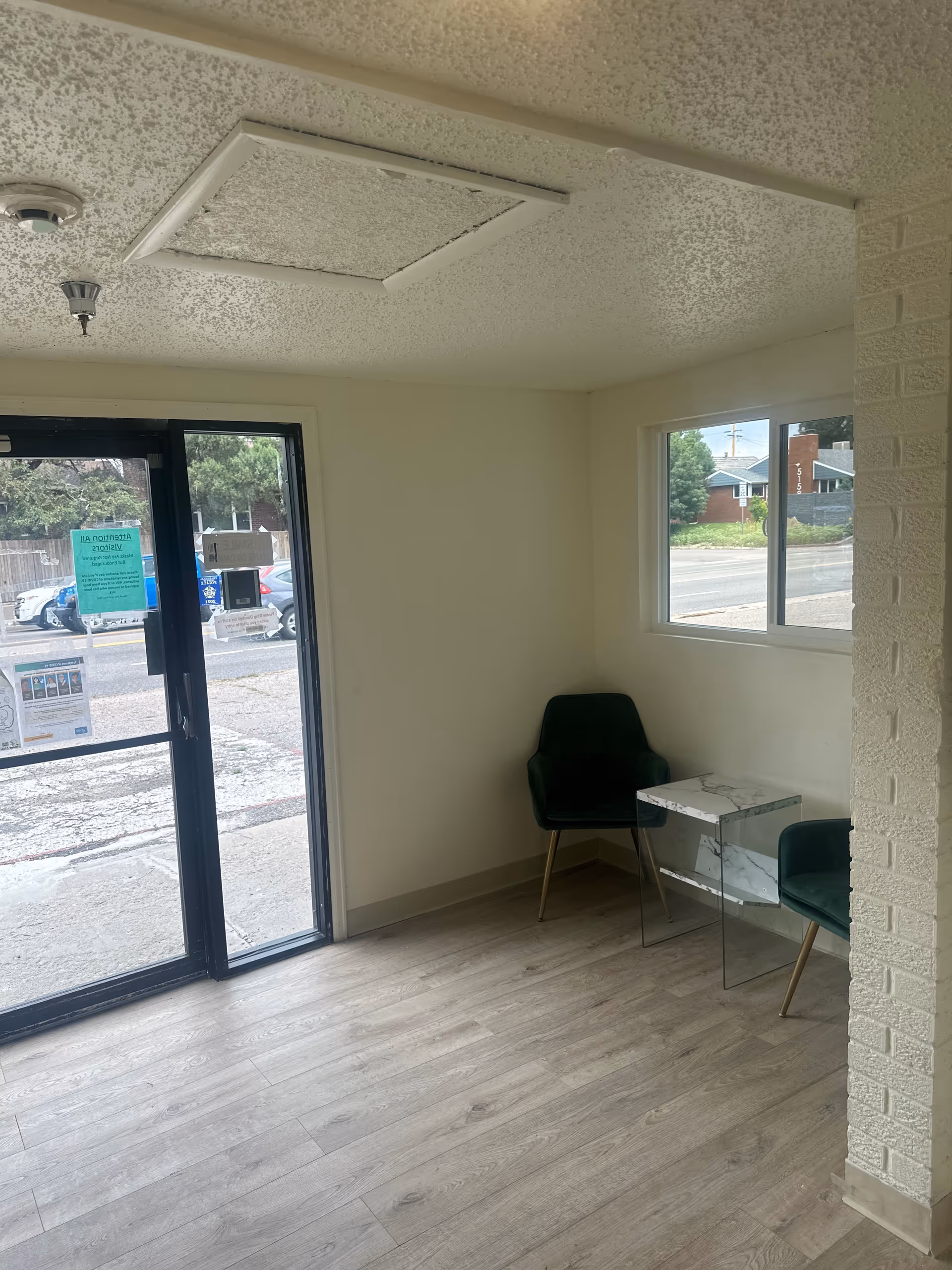 A small waiting area with two dark green chairs and a small glass and marble side table between them. The room has light-colored wooden flooring, a white textured ceiling with a ceiling vent, and a window showing a street view outside. A glass door with notices posted on it leads outside.