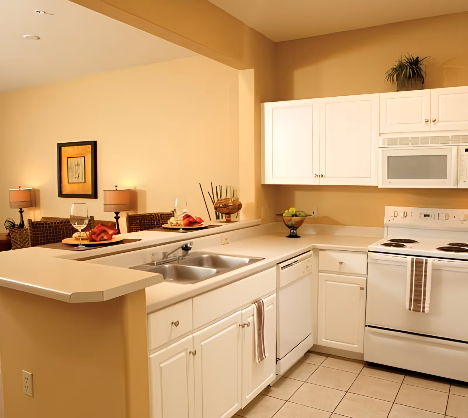 A clean and well-lit kitchen with white cabinets, a double sink, a dishwasher, an electric stove with a striped towel hanging on the handle, and a microwave above the stove. The kitchen counter extends into a breakfast bar with two place settings, each with a wine glass and red napkin. In the background, there is a dining area with wicker chairs, two table lamps, and a framed picture on the wall.