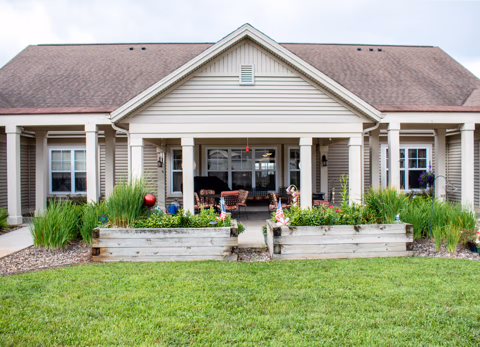 Exterior view of a single-story senior living facility building with beige siding and a brown shingled roof. The entrance area features a covered porch with several chairs and tables. In front of the porch, there are two large wooden planter boxes filled with green plants and small American flags. The lawn in front is well-maintained and green.