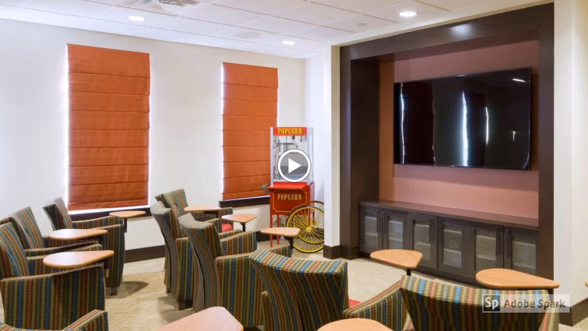 A small theater-style room with several striped upholstered chairs arranged in rows facing a large flat-screen TV mounted on a wall. There are small wooden tables attached to some chairs. Two windows with orange Roman shades are on the left wall, and a vintage-style popcorn machine is positioned near the windows.