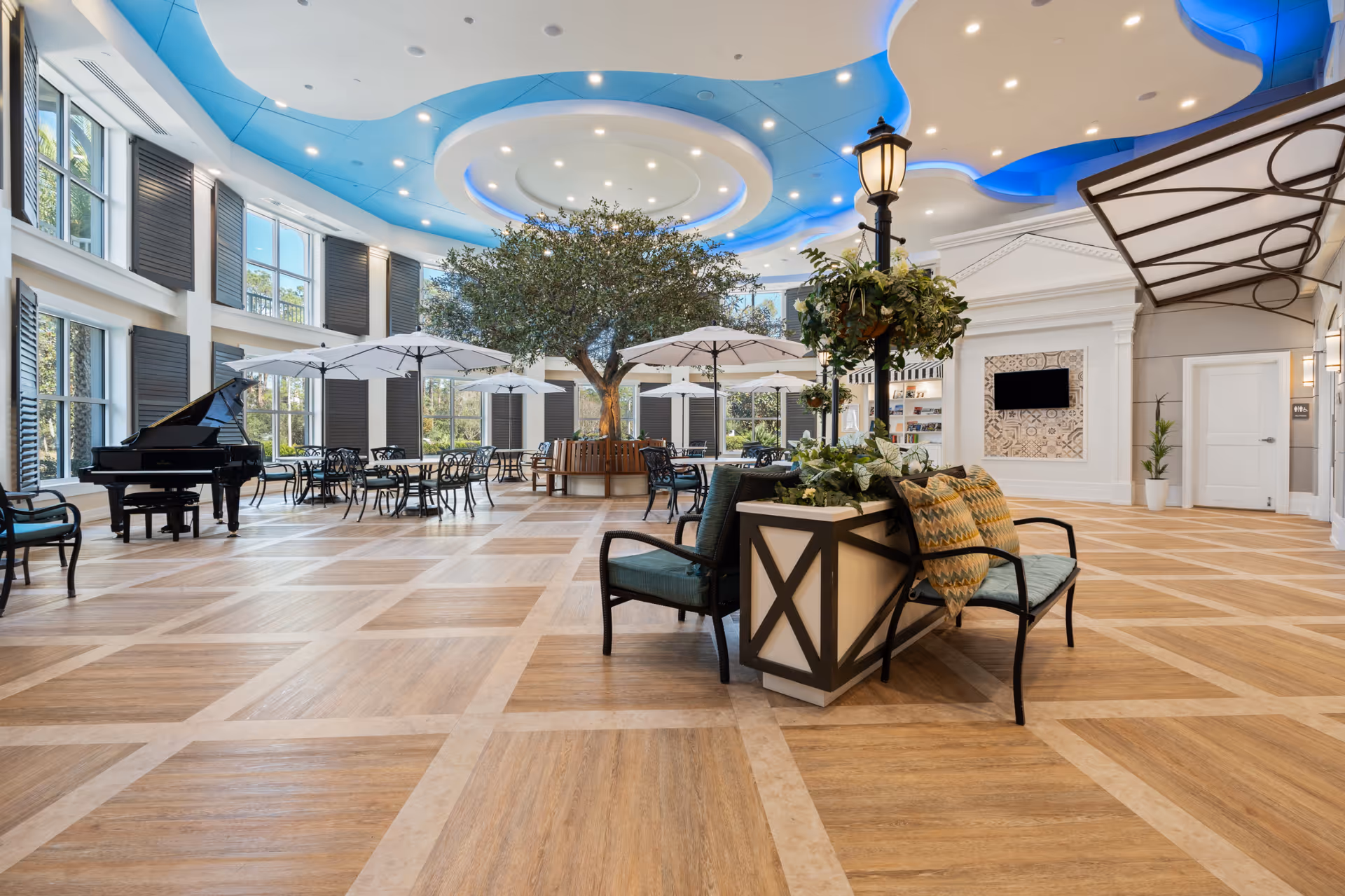 Spacious and bright common area with large windows, a grand piano, multiple tables with umbrellas, a central tree surrounded by seating, decorative ceiling lighting with blue accents, and comfortable chairs with cushions around a planter with a lamp post and greenery.