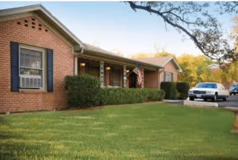 Single-story brick building with a covered porch, green bushes along the front, an American flag hanging near the entrance, and a car parked on the driveway. The scene is set on a clear day with trees in the background.